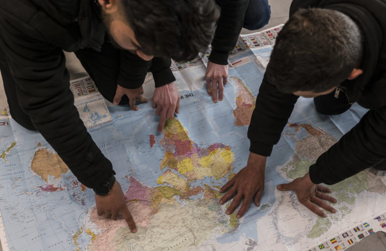 Refugees selected for resettlement to the Netherlands via the EUAA RSF take part in a Pre-Departure Orientation class, with three refugees pointing to locations on a world map placed on the floor